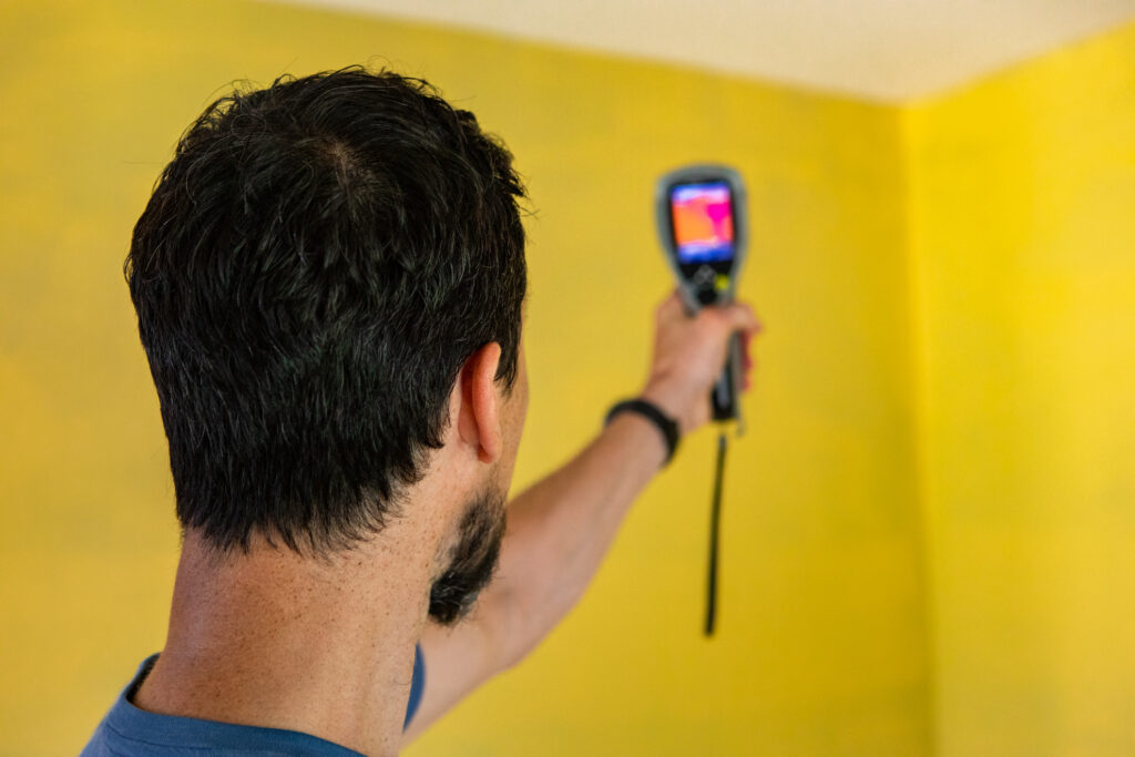 A closeup view of a home energy assessment professional using an infrared thermal imagery machine against bright yellow walls in a home.